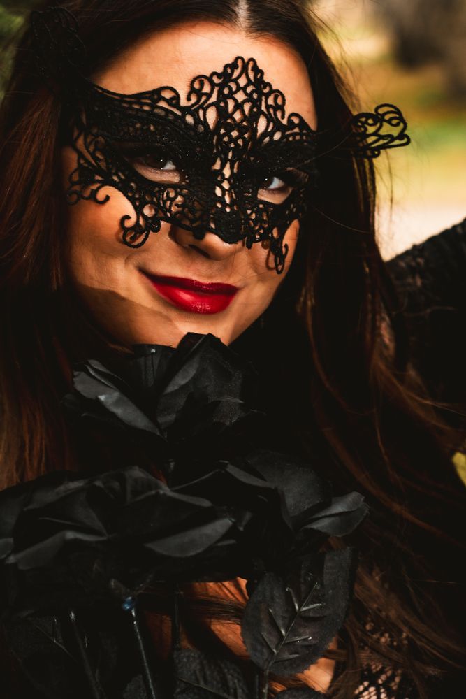 Close-up shot of ornate black lace masquerade mask with red lipstick against outdoor setting.