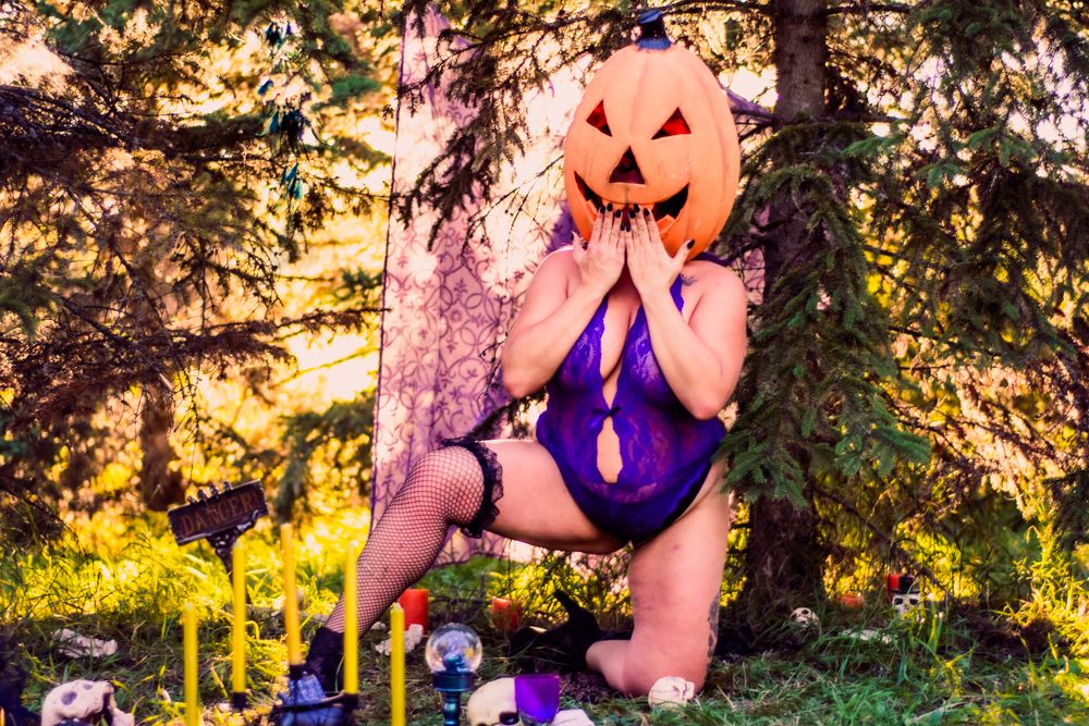 Atmospheric Halloween photo with jack-o-lantern mask and purple lingerie against forest backdrop.