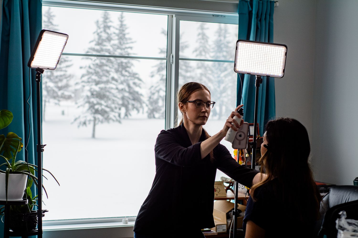 Professional makeup artist applies cosmetics in a bright room with snowy winter scenery visible through window.
