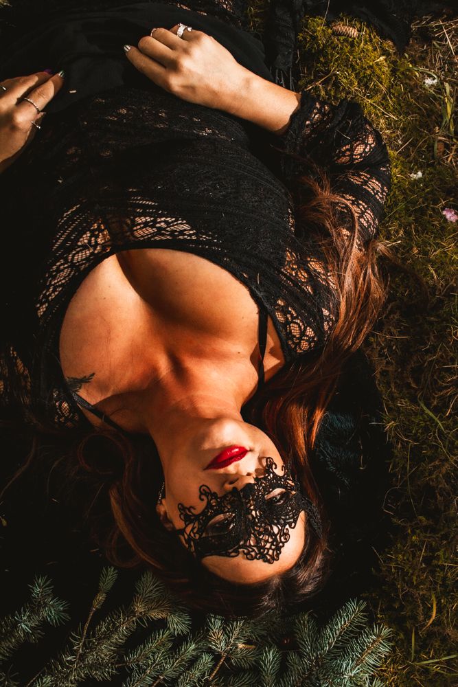 Overhead view of figure in black lace mask lying on mossy ground with white candle nearby.