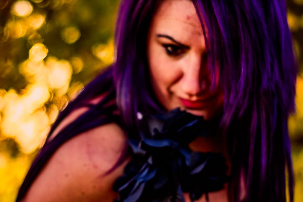 Artistic portrait with vibrant purple hair and dark blue roses against golden bokeh lights.