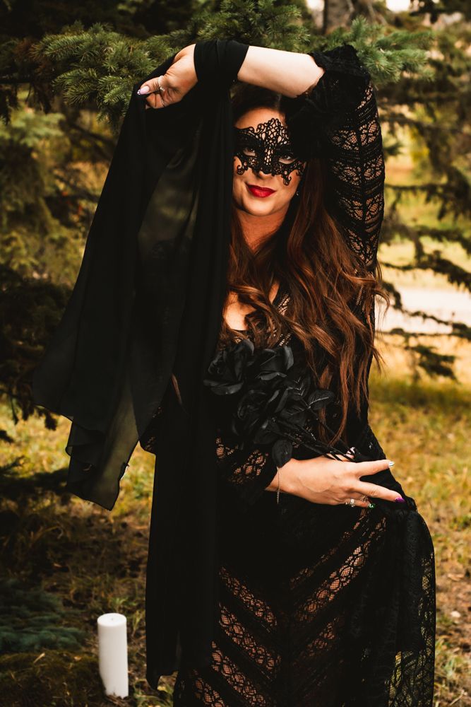 Dramatic pose in flowing black lace dress and masquerade mask outdoors near evergreen trees.