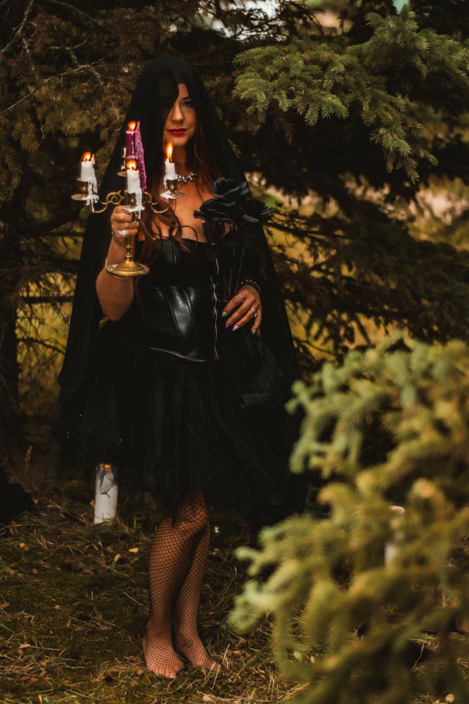 Gothic styled photoshoot with black dress and candelabra among evergreen trees.