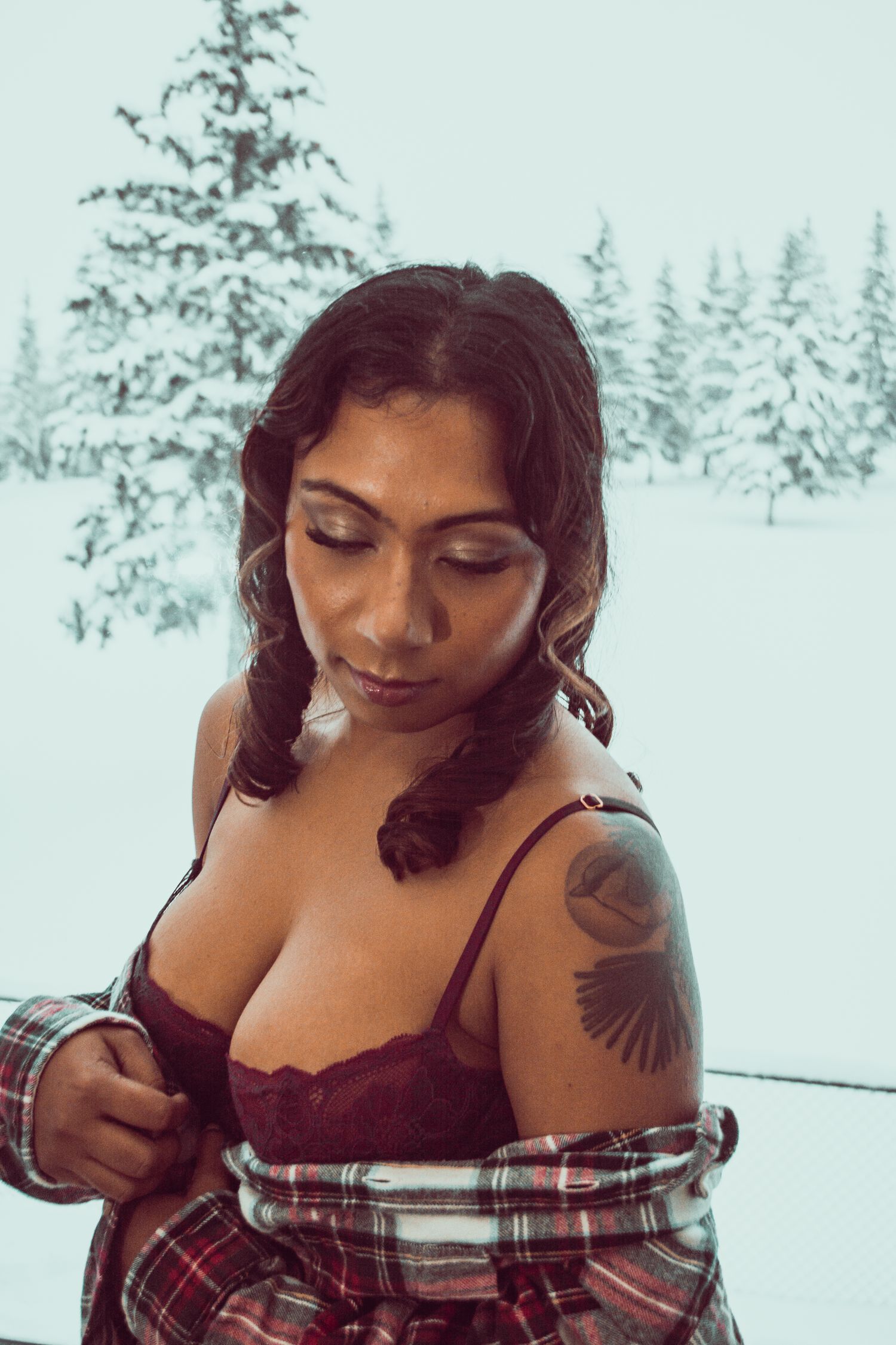 Person in burgundy lingerie and plaid shirt poses by window with snowy forest backdrop.