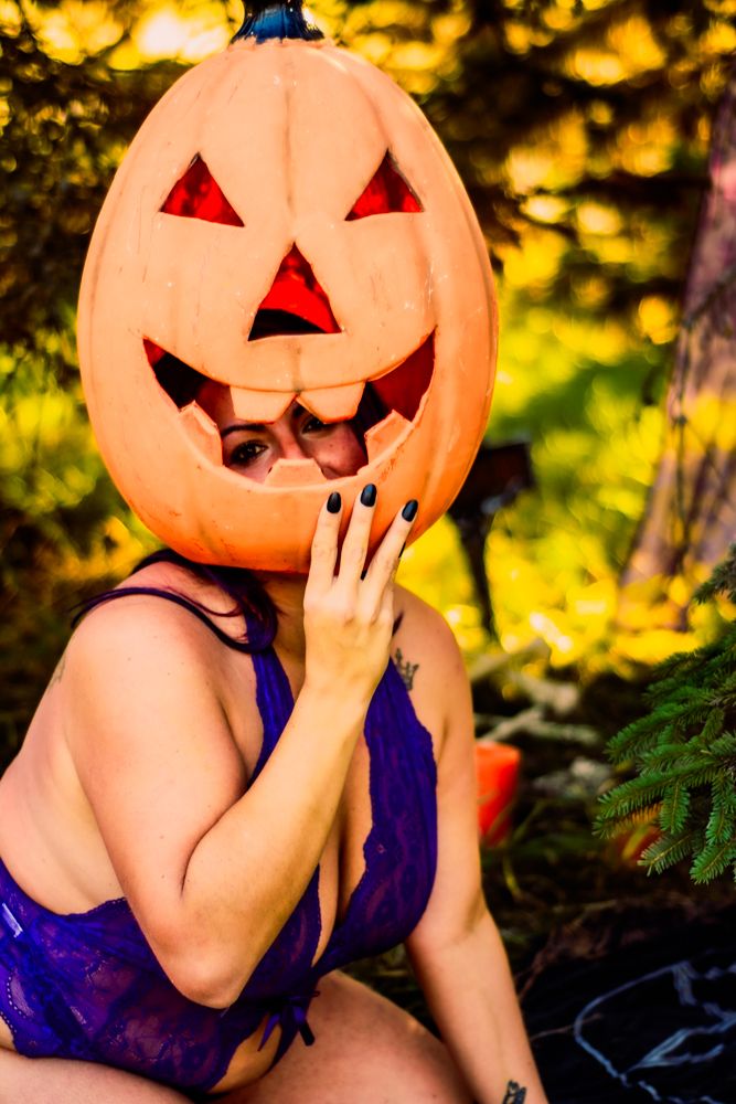 Model wearing a giant jack-o'-lantern head and purple swimsuit poses in a golden-lit outdoor setting.