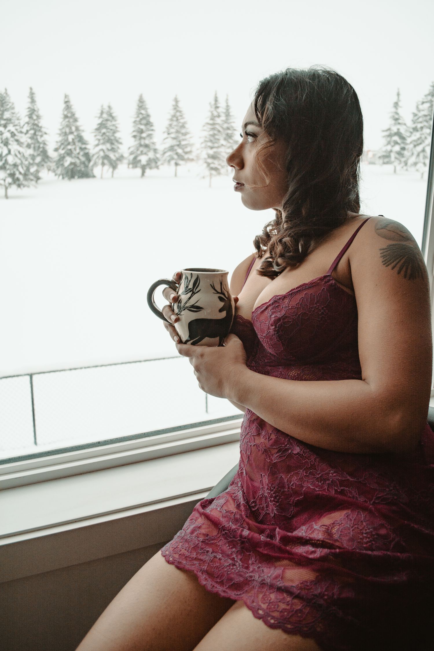 Woman in burgundy lace holding decorative mug while looking out at snowy scene.