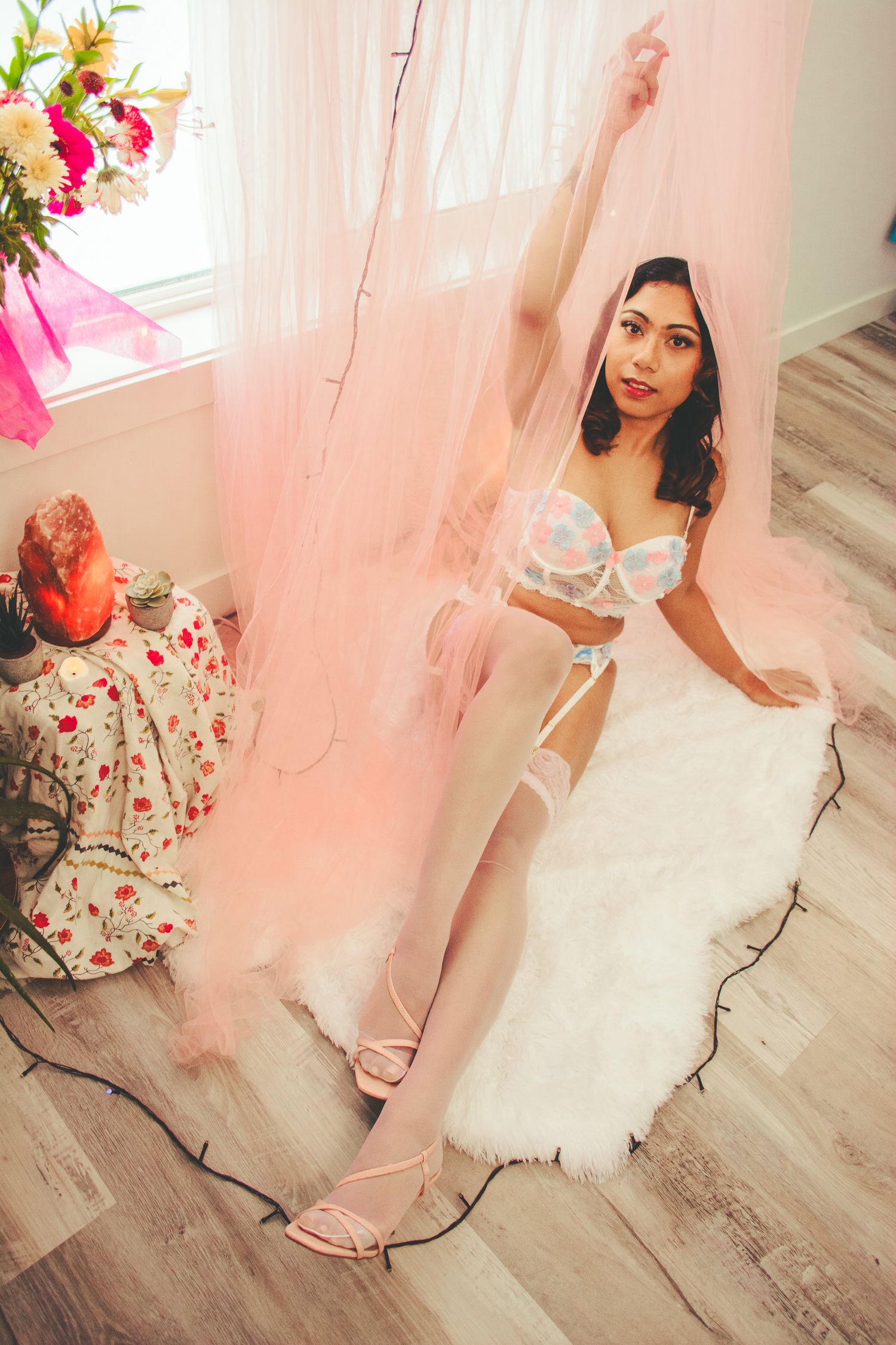 Artistic boudoir shot with pink sheer curtains, floral arrangements, and white fur rug.