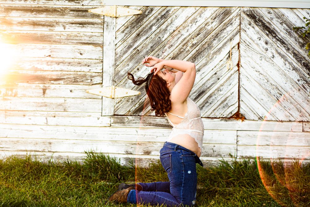 Dynamic pose against rustic barn wall with white lace top and denim in afternoon light.