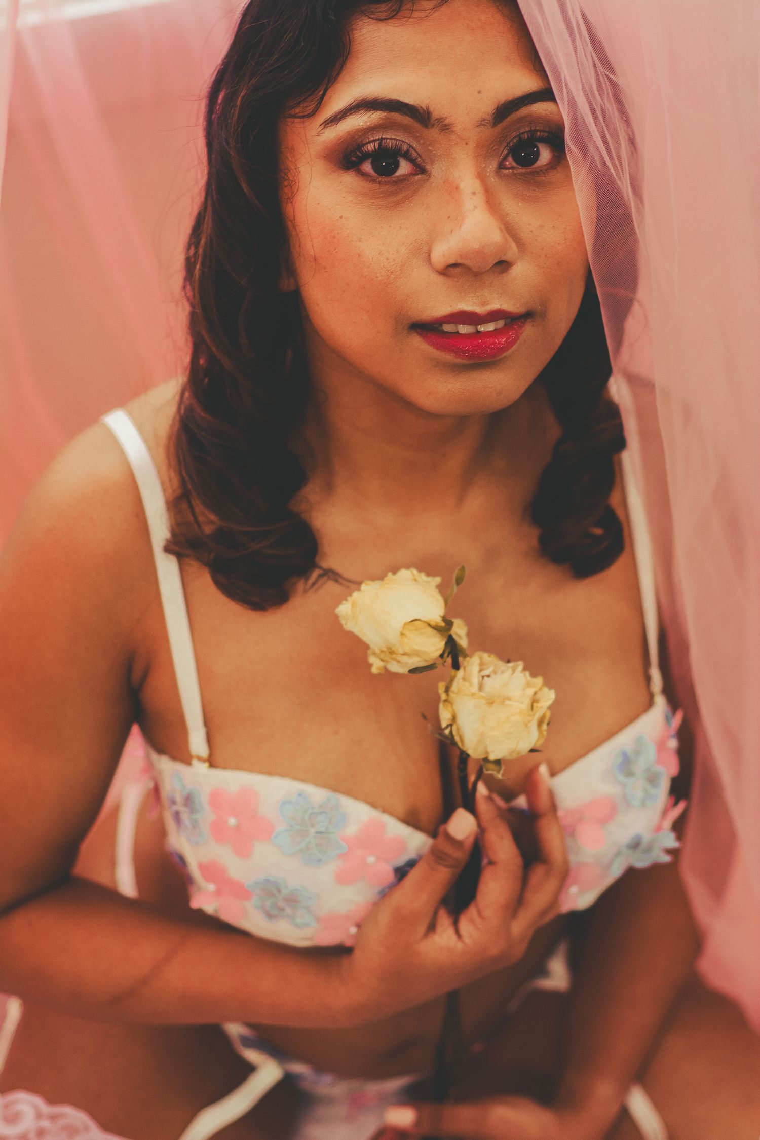 Close-up boudoir portrait with cream roses and floral lingerie under pink veil.