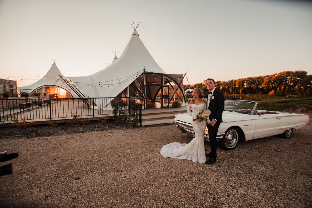 Classic white convertible car parked in front of wedding tent at sunset.