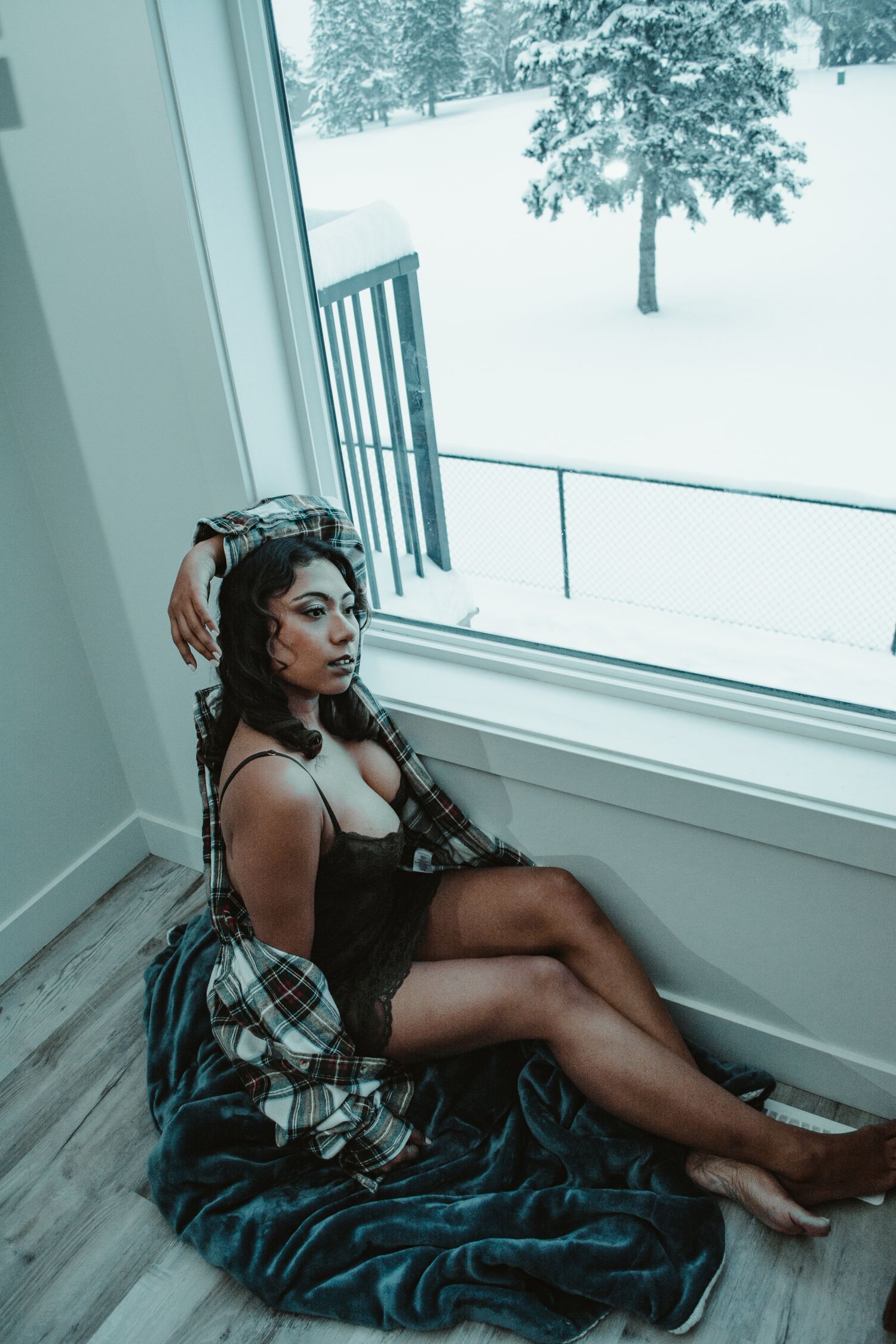 Woman in lingerie lounges by snowy window with plaid flannel and blue blanket in moody indoor setting.