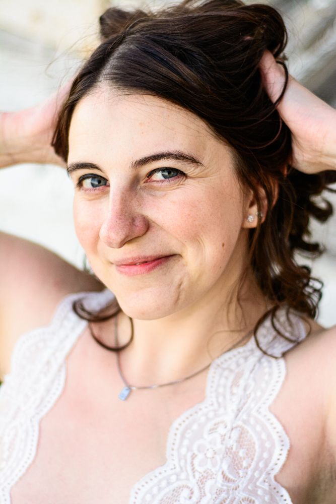 Close-up portrait in white lace showing natural smile and soft curls against weathered wall.