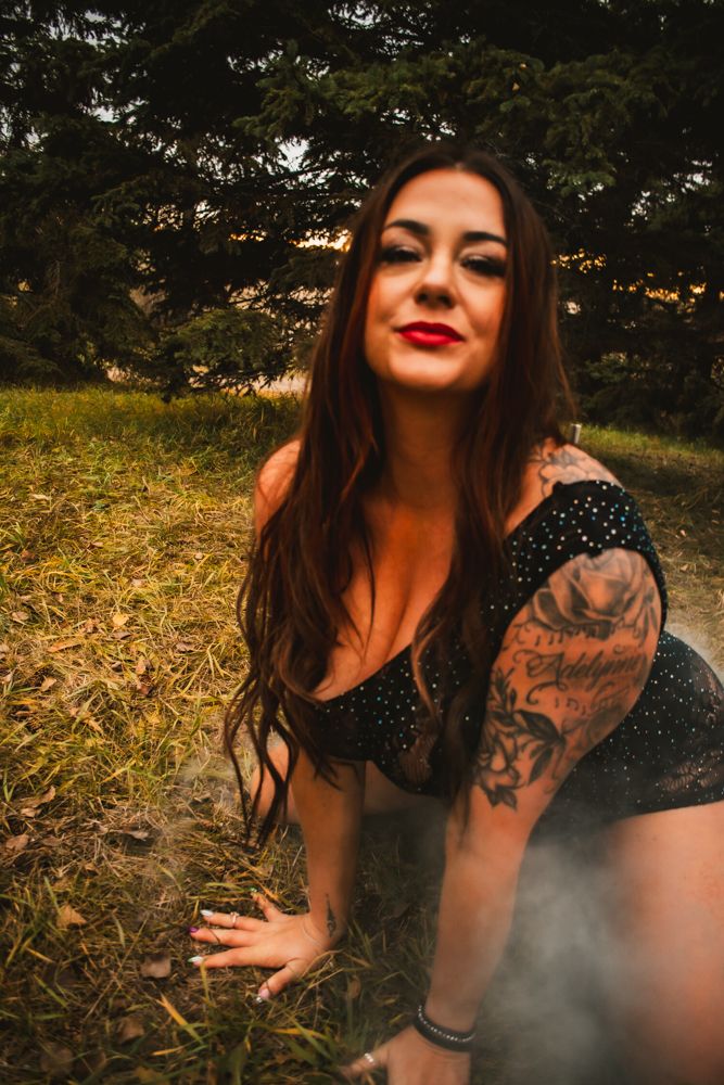 Portrait outdoors showing long dark hair and arm tattoo against wooded background with atmospheric lighting.