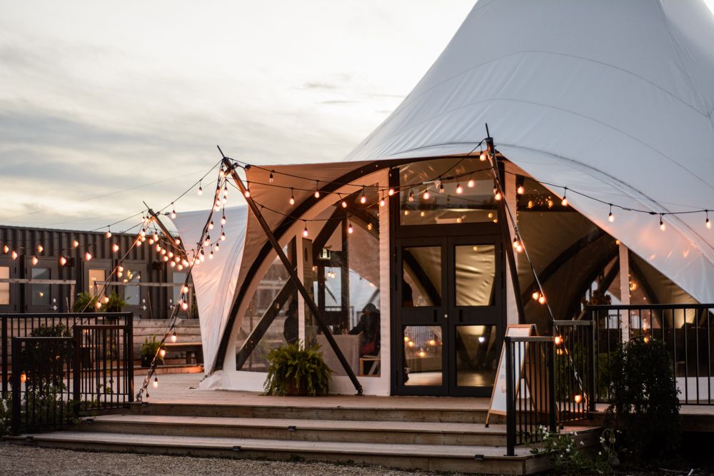 Outdoor wedding tent pavilion decorated with string lights at dusk against cloudy sky.