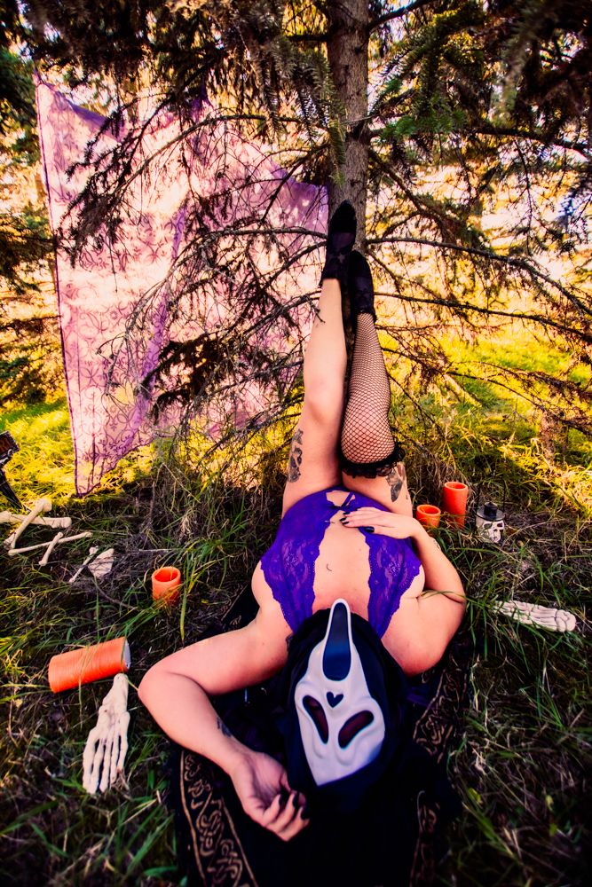 Halloween themed photo with purple lingerie and ghost mask in forest with spooky candles at sunset.