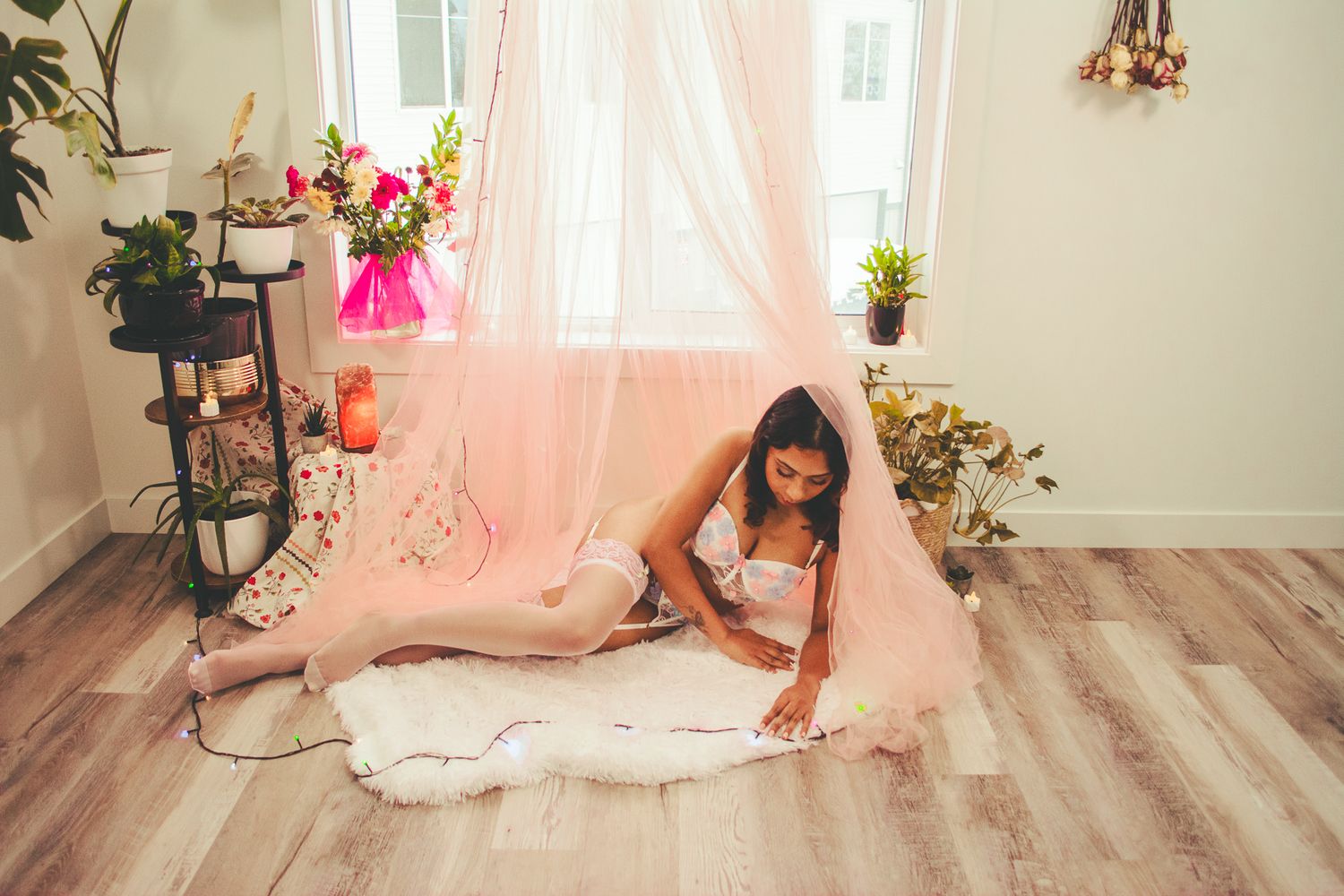 Full length boudoir portrait on wooden floor with pink curtains and potted plants.