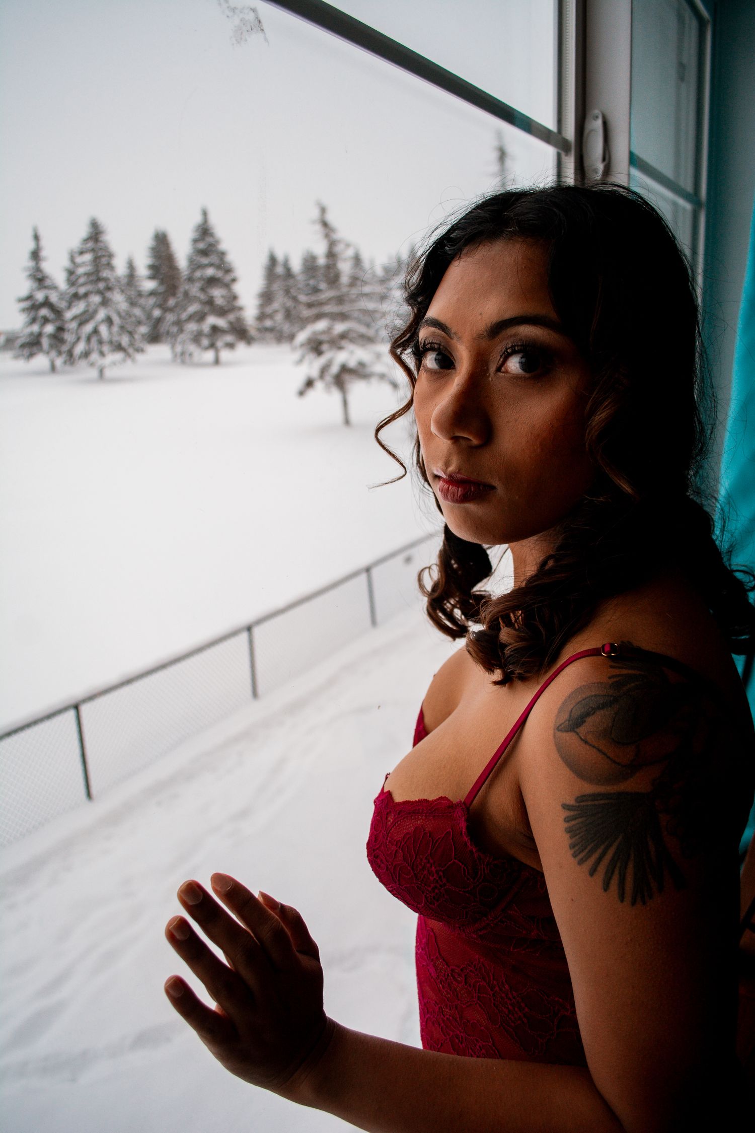 Woman in red lingerie poses by window overlooking snow-covered pine trees.