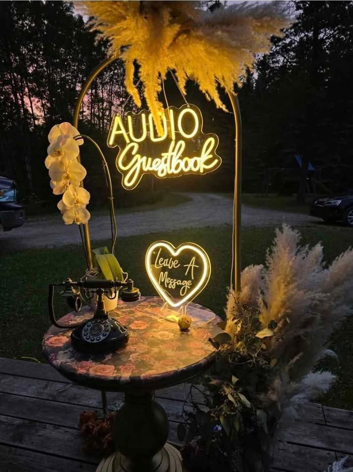 A romantic outdoor setup with illuminated neon signs and pampas grass decorations on a rustic wooden table at dusk.