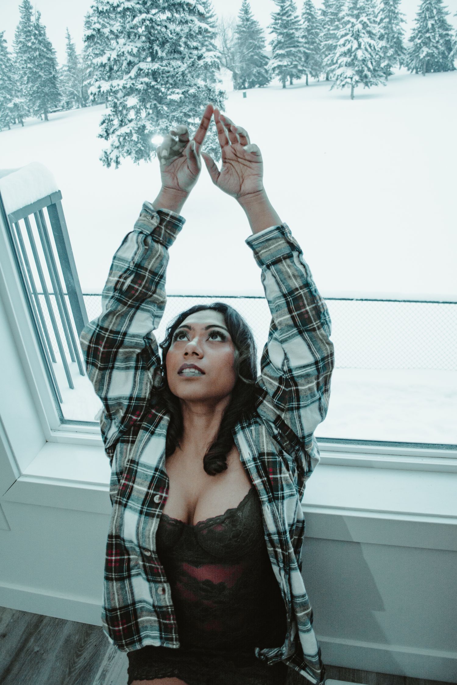 Person in plaid flannel reaching up toward window with snowy forest view.