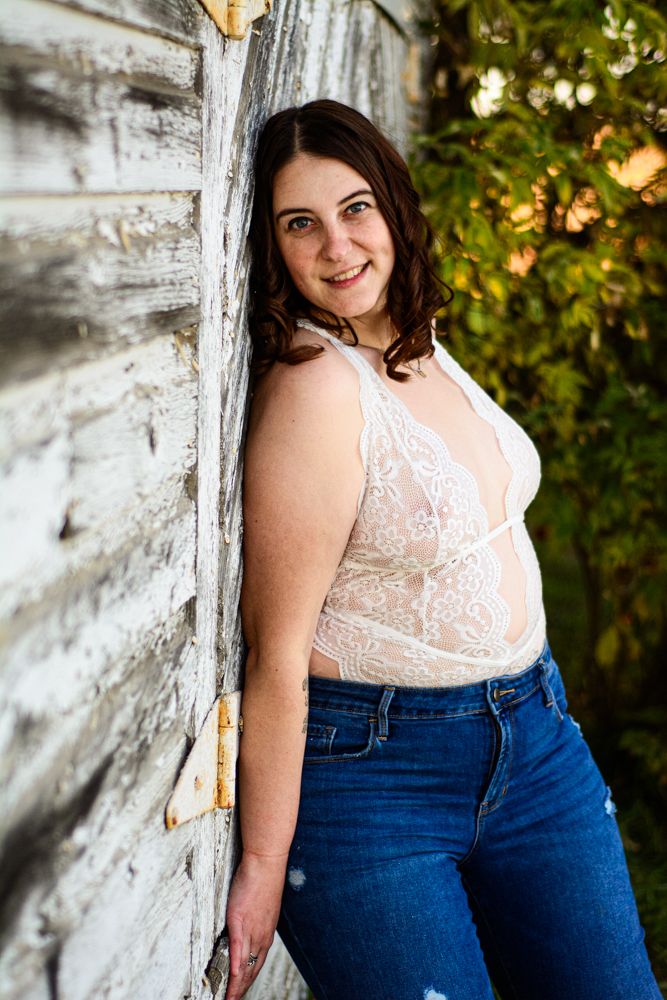 Portrait against weathered wood wall wearing white lace bodysuit and jeans in natural lighting.