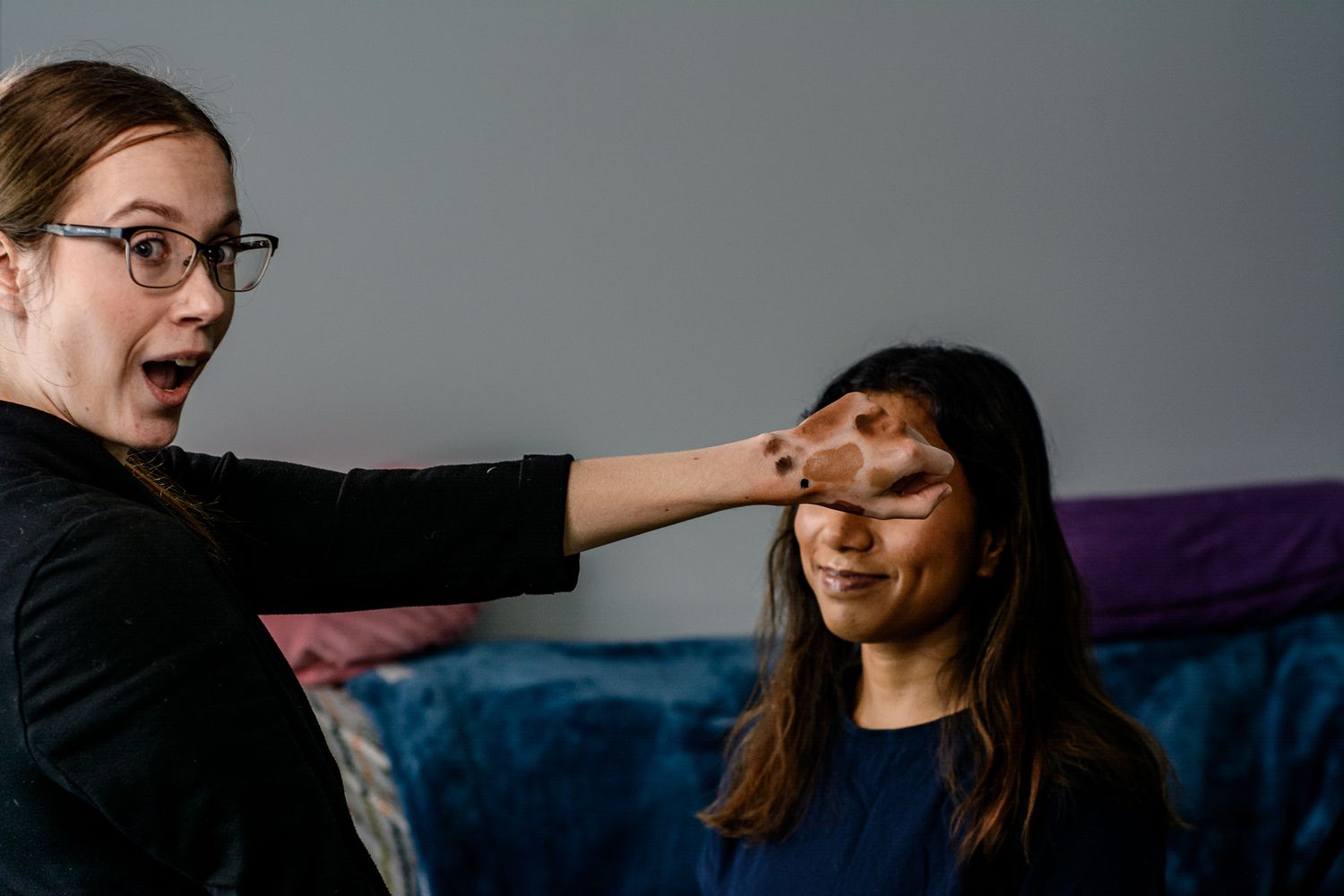 Makeup artist wearing glasses gestures playfully while working with client during beauty session.