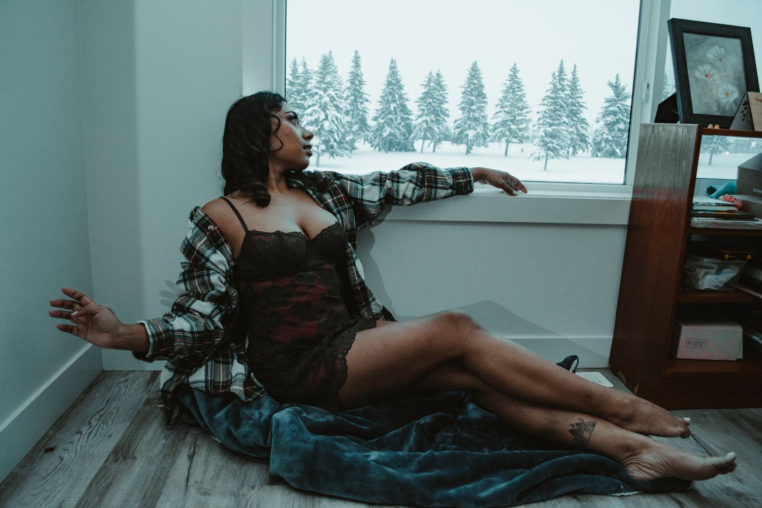 Woman in lingerie poses on floor with snowy evergreen forest visible through window.