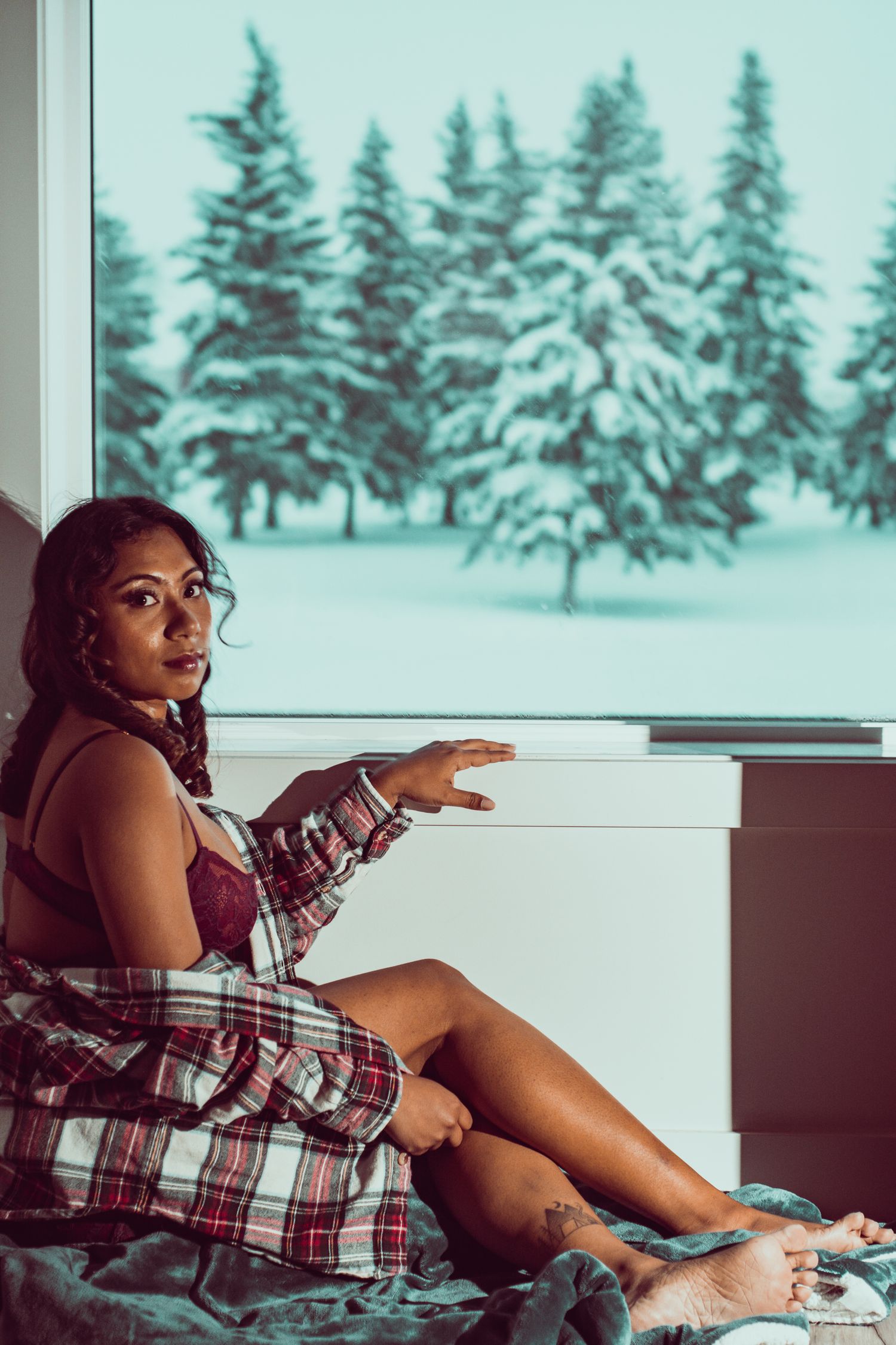 Model lounges by window overlooking snow-covered pine trees in intimate photoshoot setting.