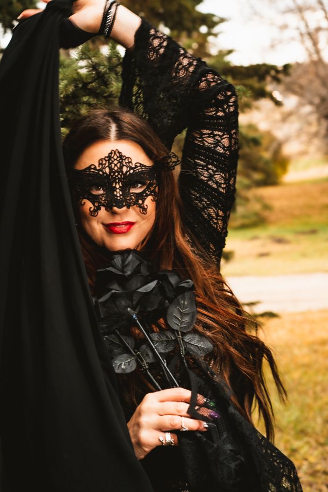 Figure in black lace masquerade mask and gothic dress poses dramatically against natural outdoor backdrop.