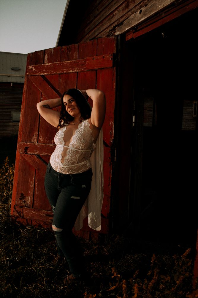 Figure in lace top posing against weathered red barn door at sunset.