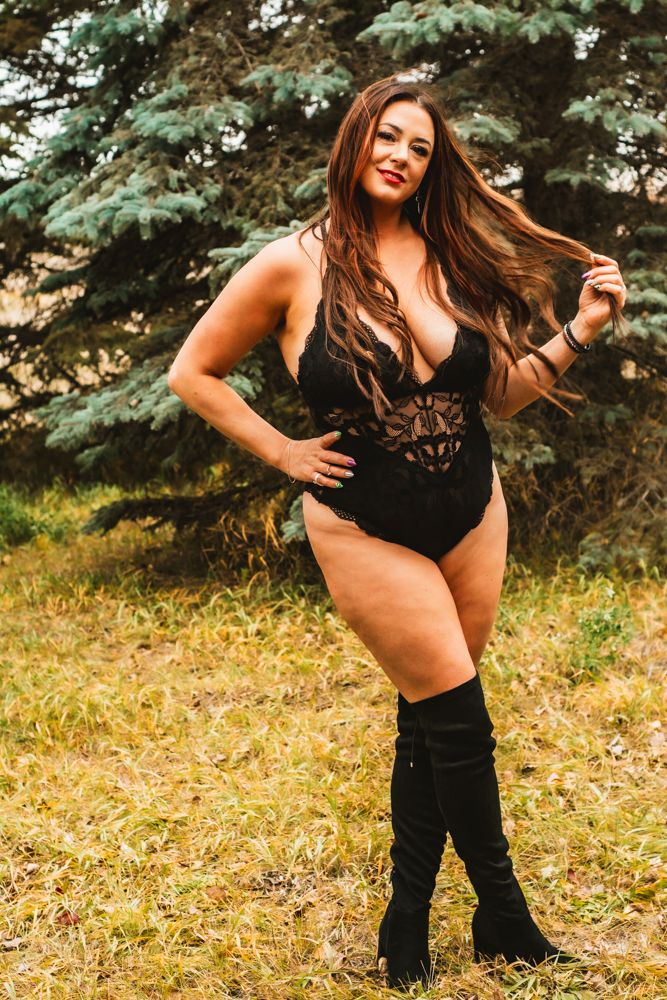 Body-positive portrait showing black lace bodysuit and knee-high boots in outdoor forest setting.