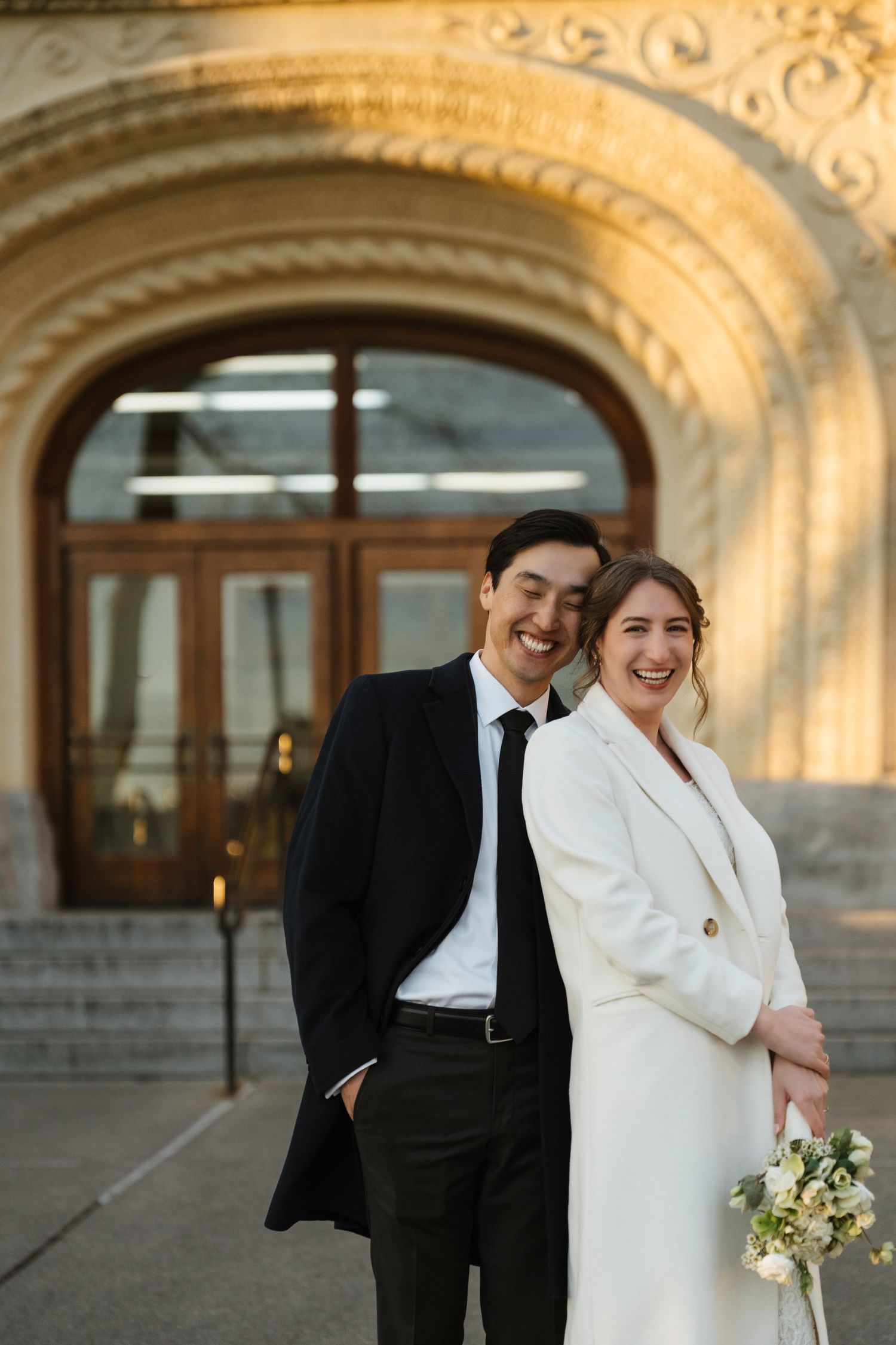 Smiling bride in white coat holding bouquet with groom in black suit posing before ornate stone building entrance.
