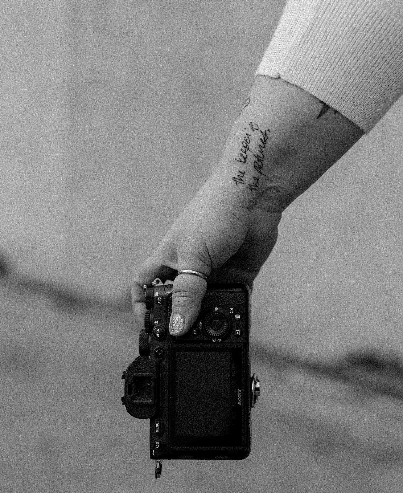 A black and white photo shows a hand holding a camera with a small tattoo visible on the wrist.