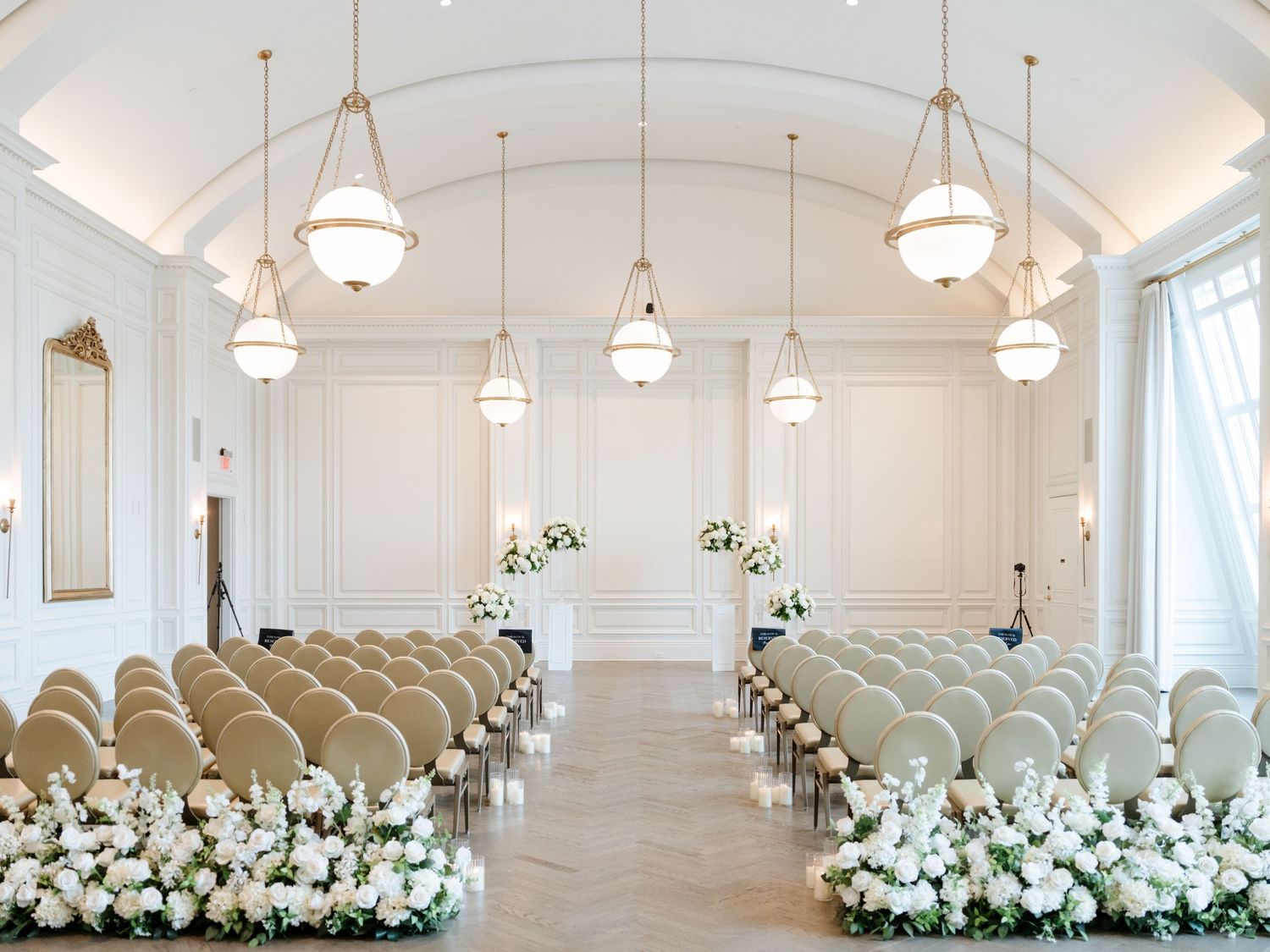Elegant white wedding ceremony setup in a bright room with hanging pendant lights and white floral arrangements lining the aisle.
