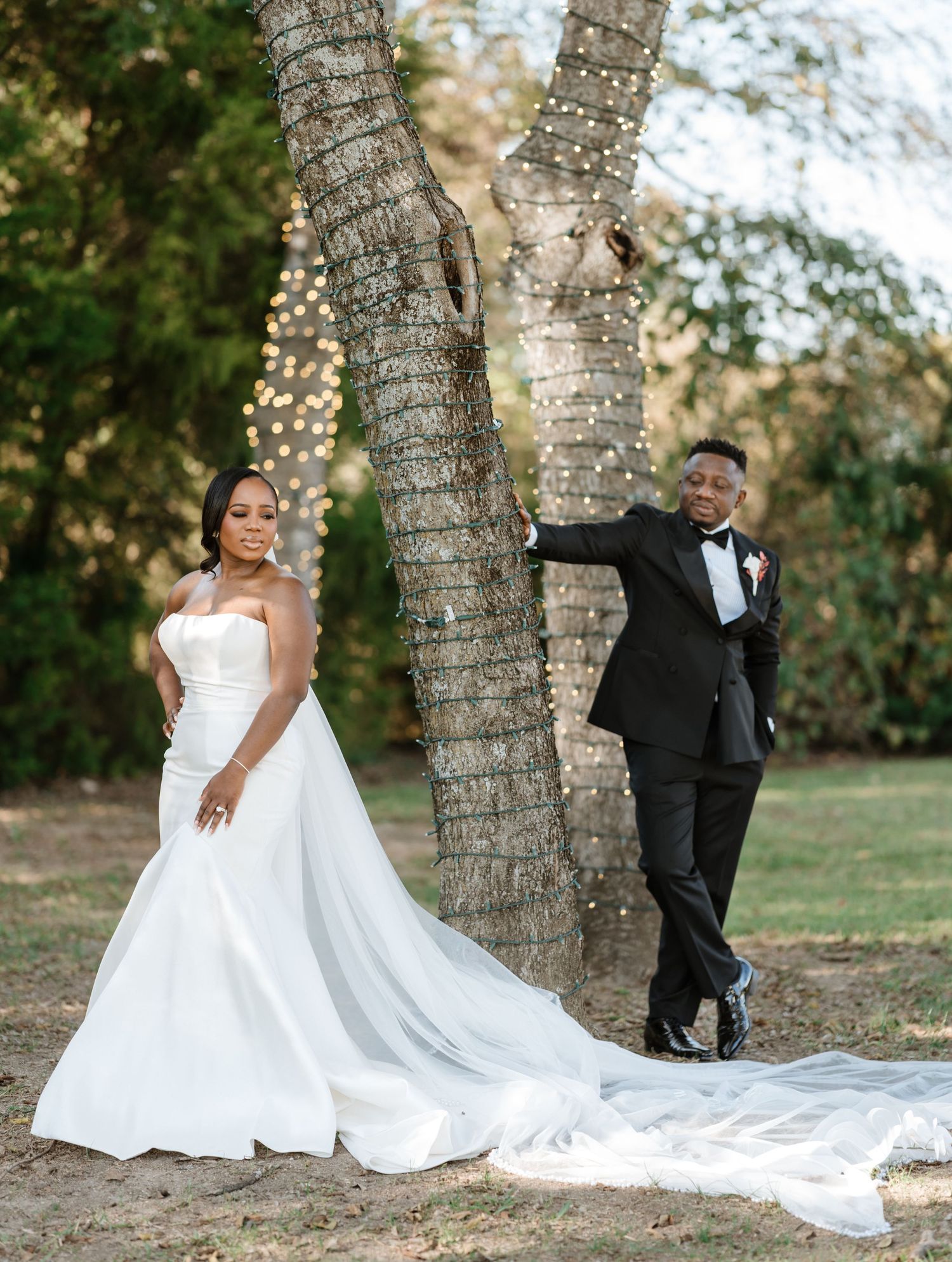 A romantic wedding couple poses playfully between palm trees in an outdoor garden setting.