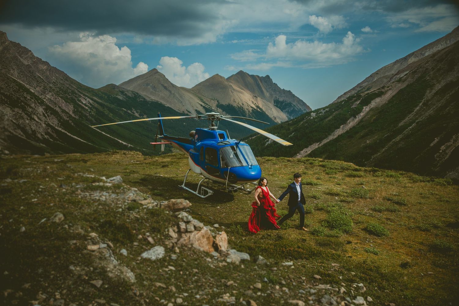 Couple in formal wear walks from a blue helicopter on a mountain meadow with rocky peaks under cloudy skies.