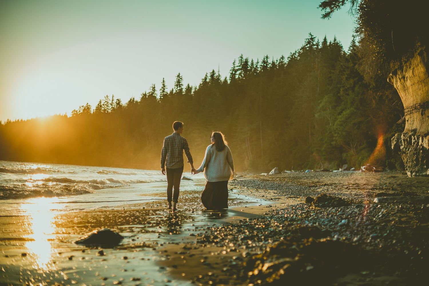 Couple holding hands walking along a rocky beach at golden sunset with misty forest backdrop.