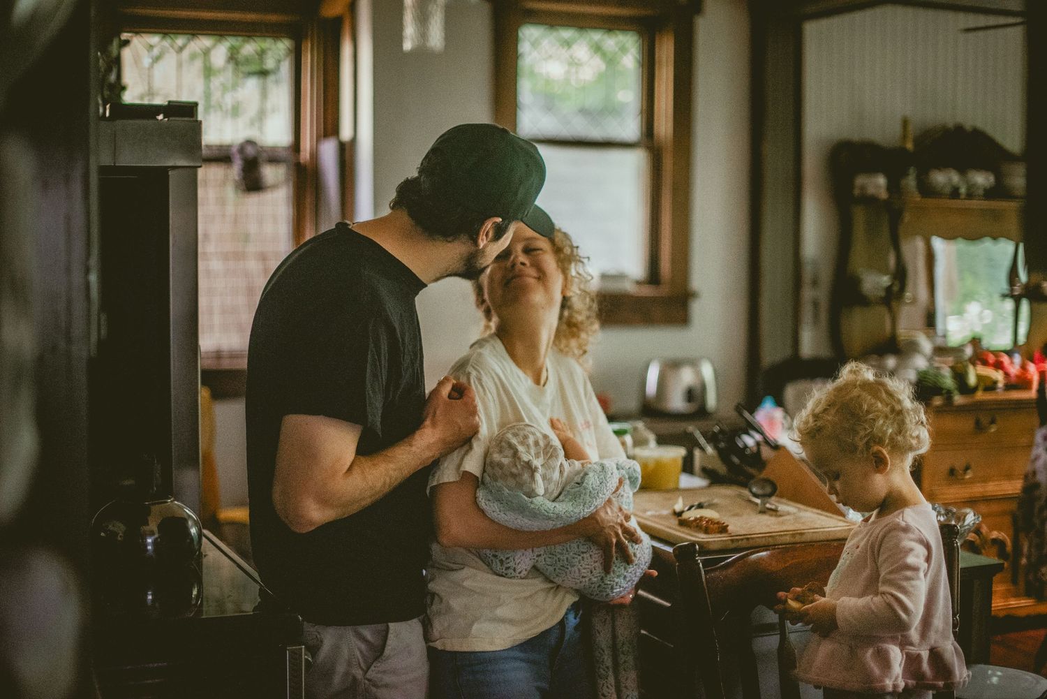 A couple shares a tender kiss while holding a baby in a rustic kitchen, as a young child plays nearby.