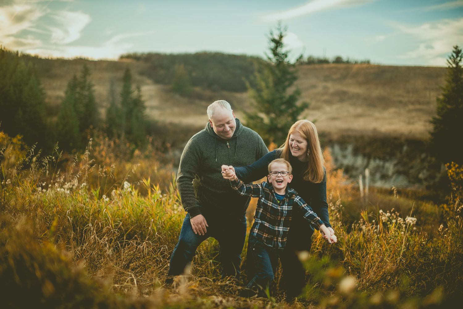 A couple embraces in a golden autumn field during sunset with rolling hills and evergreen trees in the background.