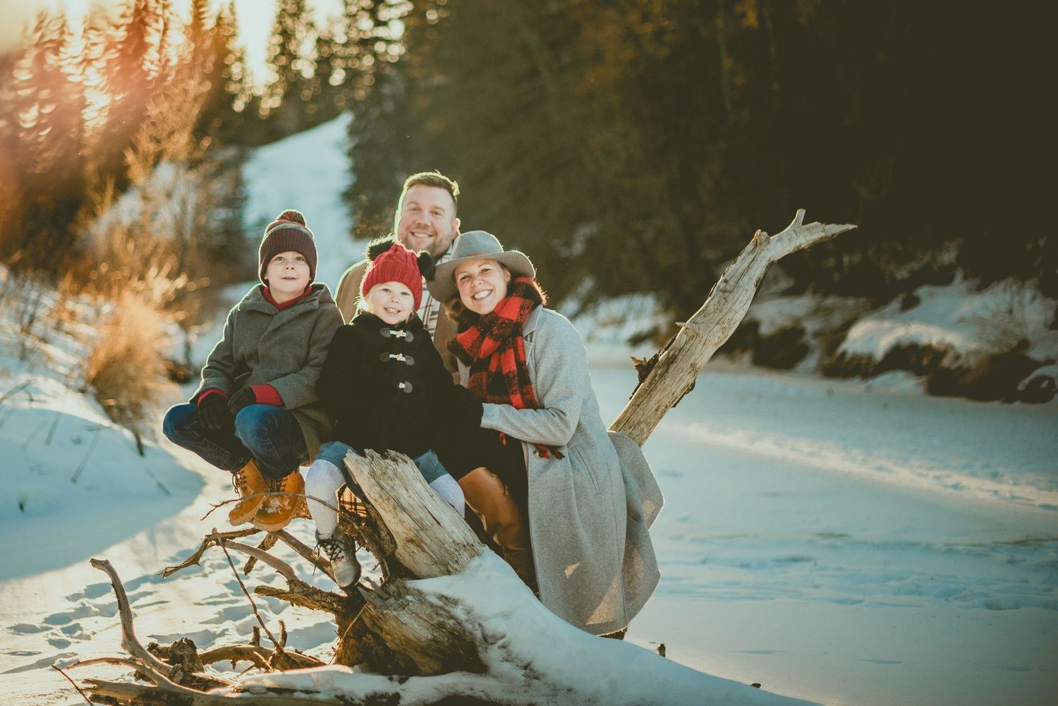 Family sits together on snow-covered log at sunset with winter forest in background during outdoor winter photography adventure.