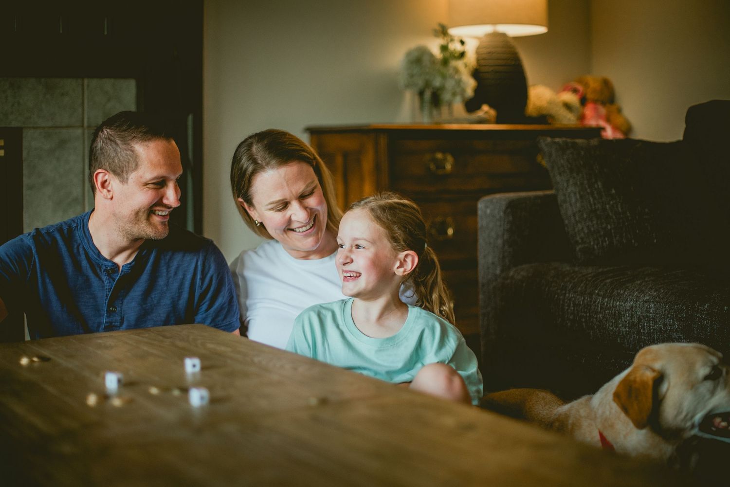 A happy family of three smiles together at a wooden table with a dog resting nearby in a cozy home.