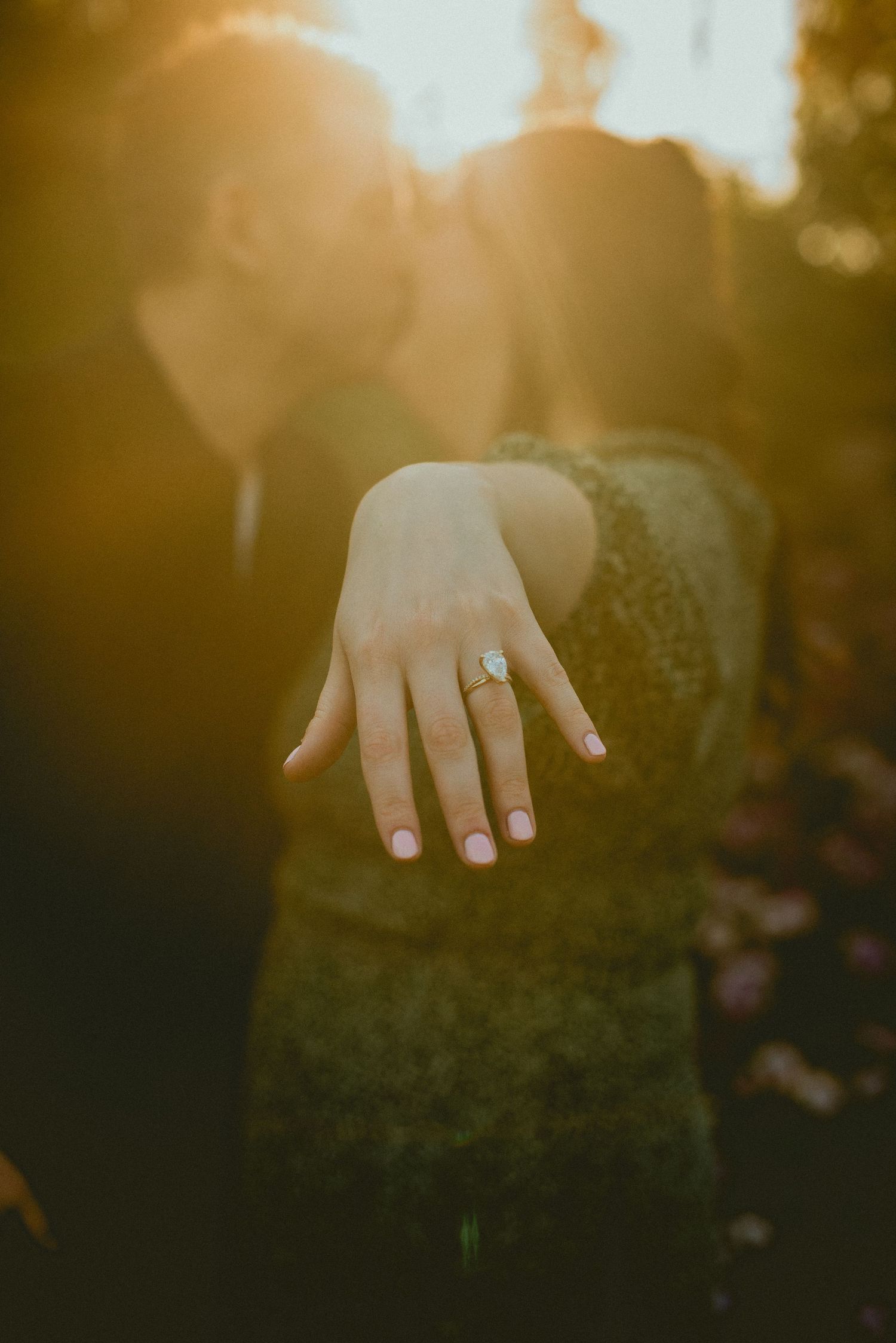 Couple kissing at sunset while showing off an elegant engagement ring with a large center stone in golden light.