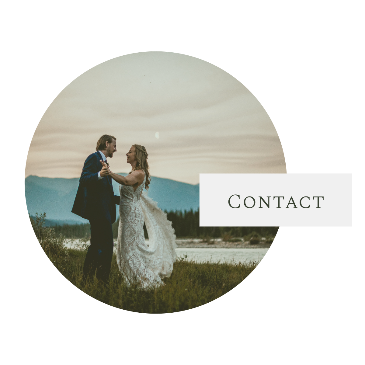 A couple in wedding attire stands together in a scenic meadow at sunset with mountains in the background.