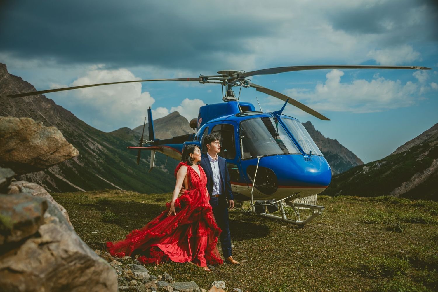 Couple in red dress poses before blue helicopter in dramatic mountain landscape with stormy skies.