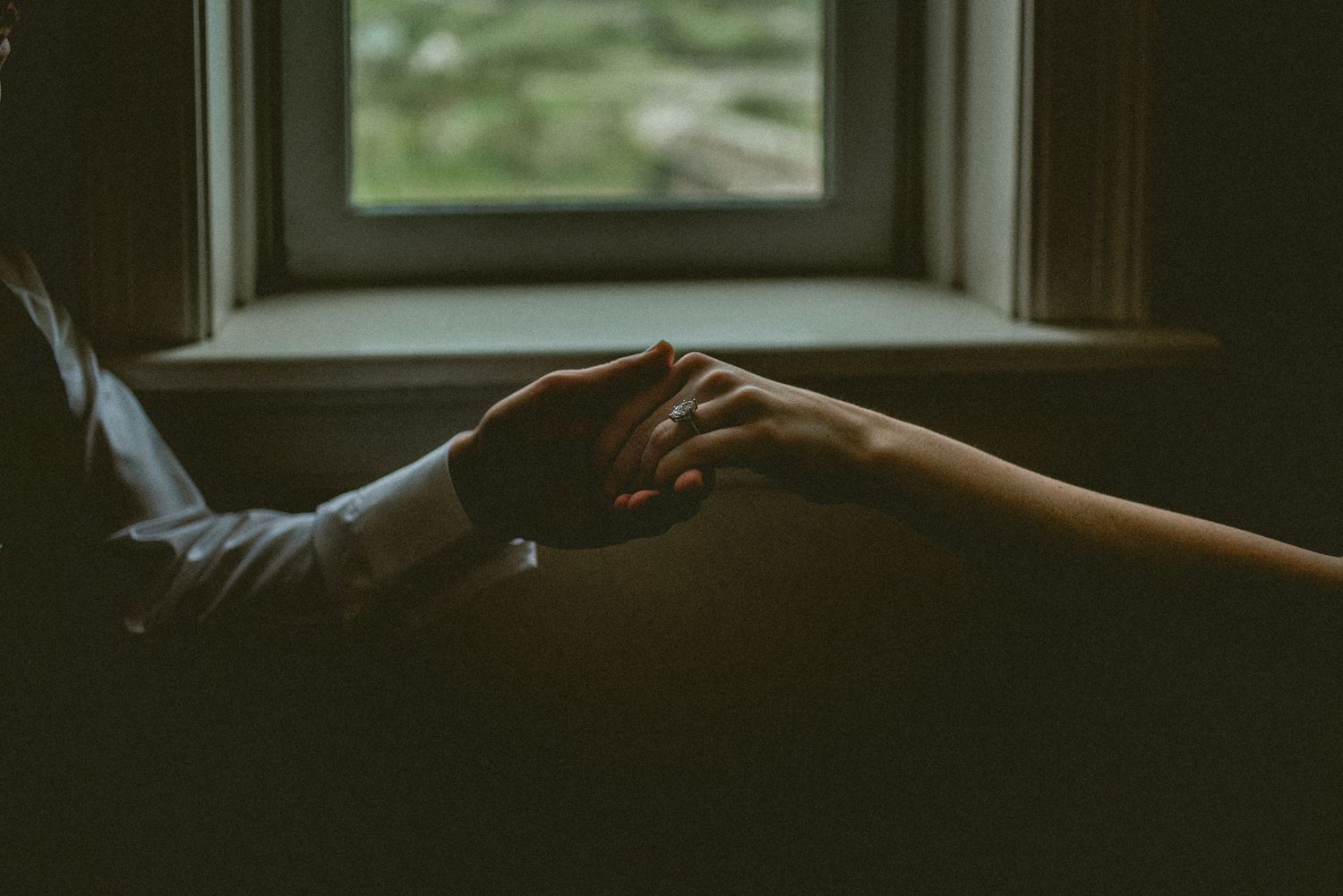 Two people holding hands tenderly near a window in soft, romantic lighting.