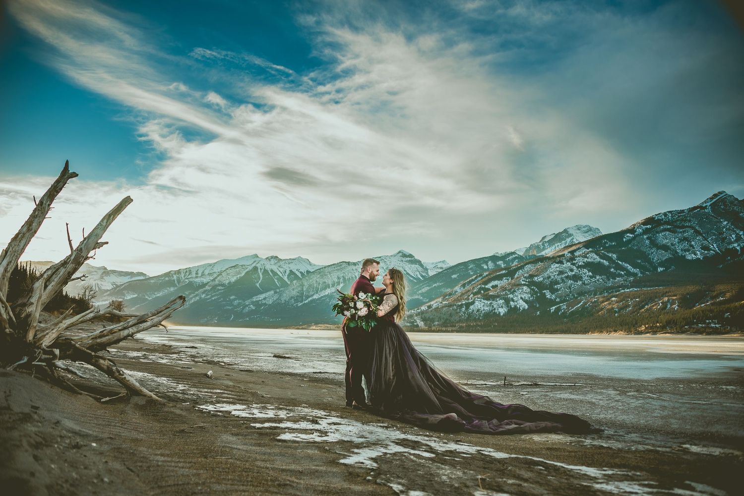 Couple in black wedding attire embracing on a mountain lakeside beach with dramatic cloudy skies and snowy peaks.