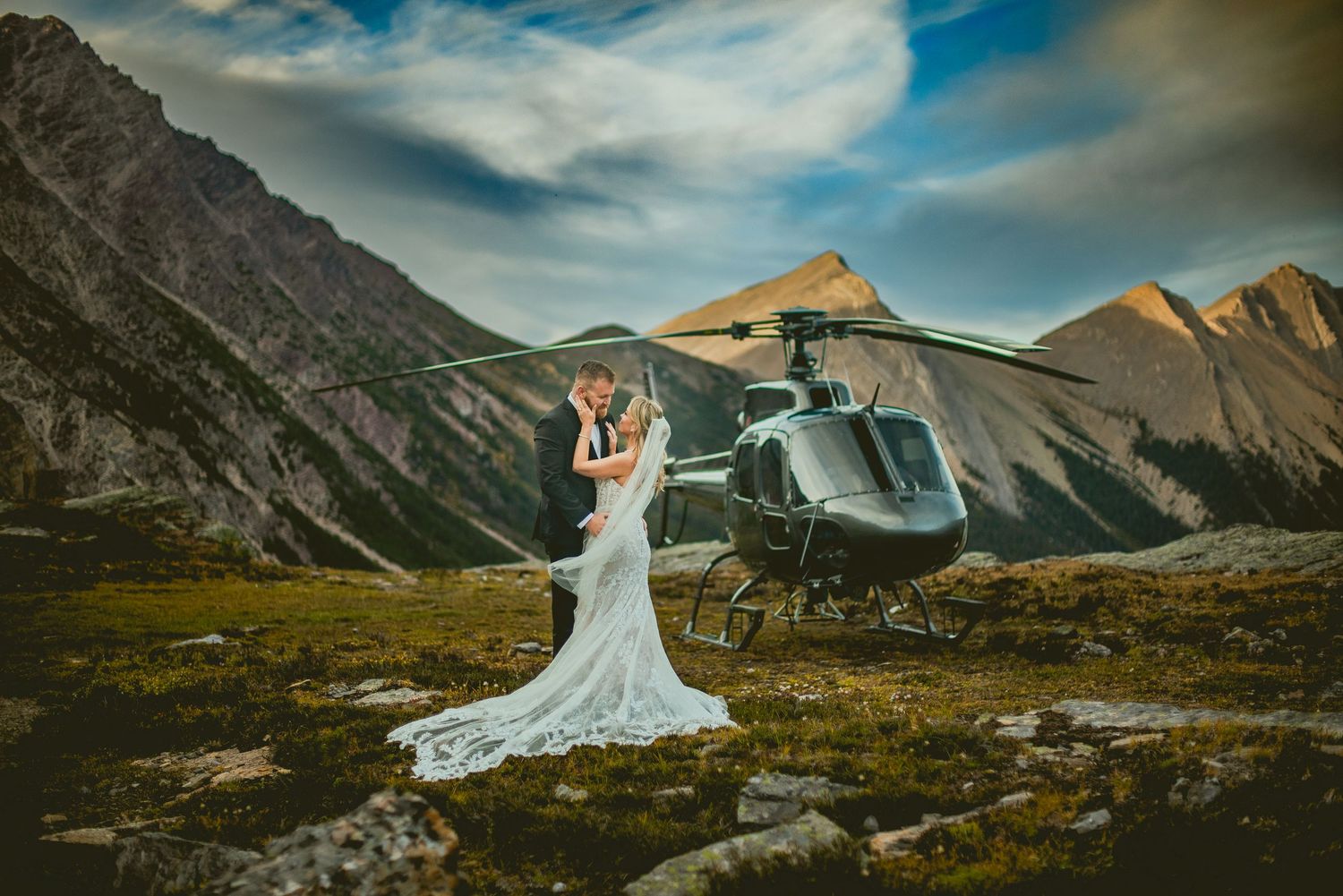 Bride in a long white gown embraces her groom beside a helicopter on a rocky mountain meadow beneath dramatic clouds and rugged peaks.