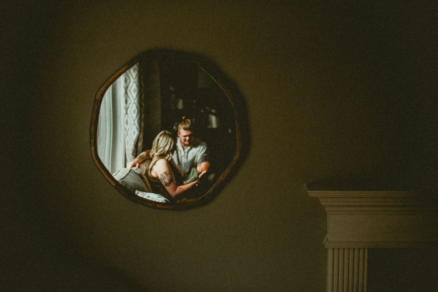 Couple reflected in an octagonal mirror on a dark wall, sharing an intimate moment near a bright window.