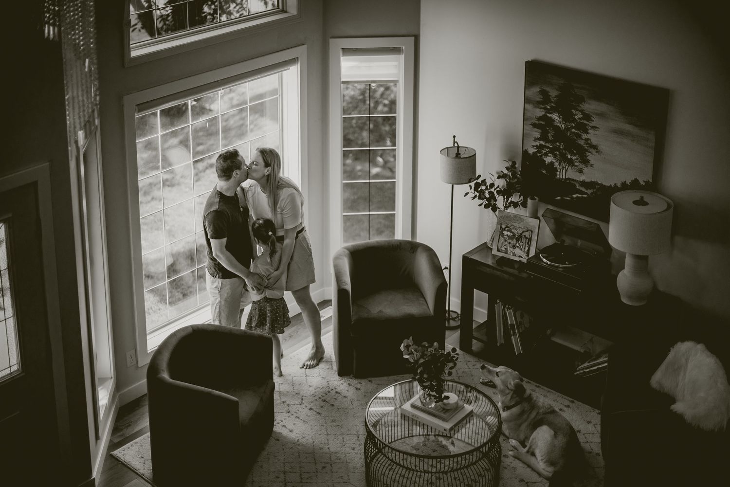 Black and white photo of a couple sharing a tender moment by large windows in a cozy living room.