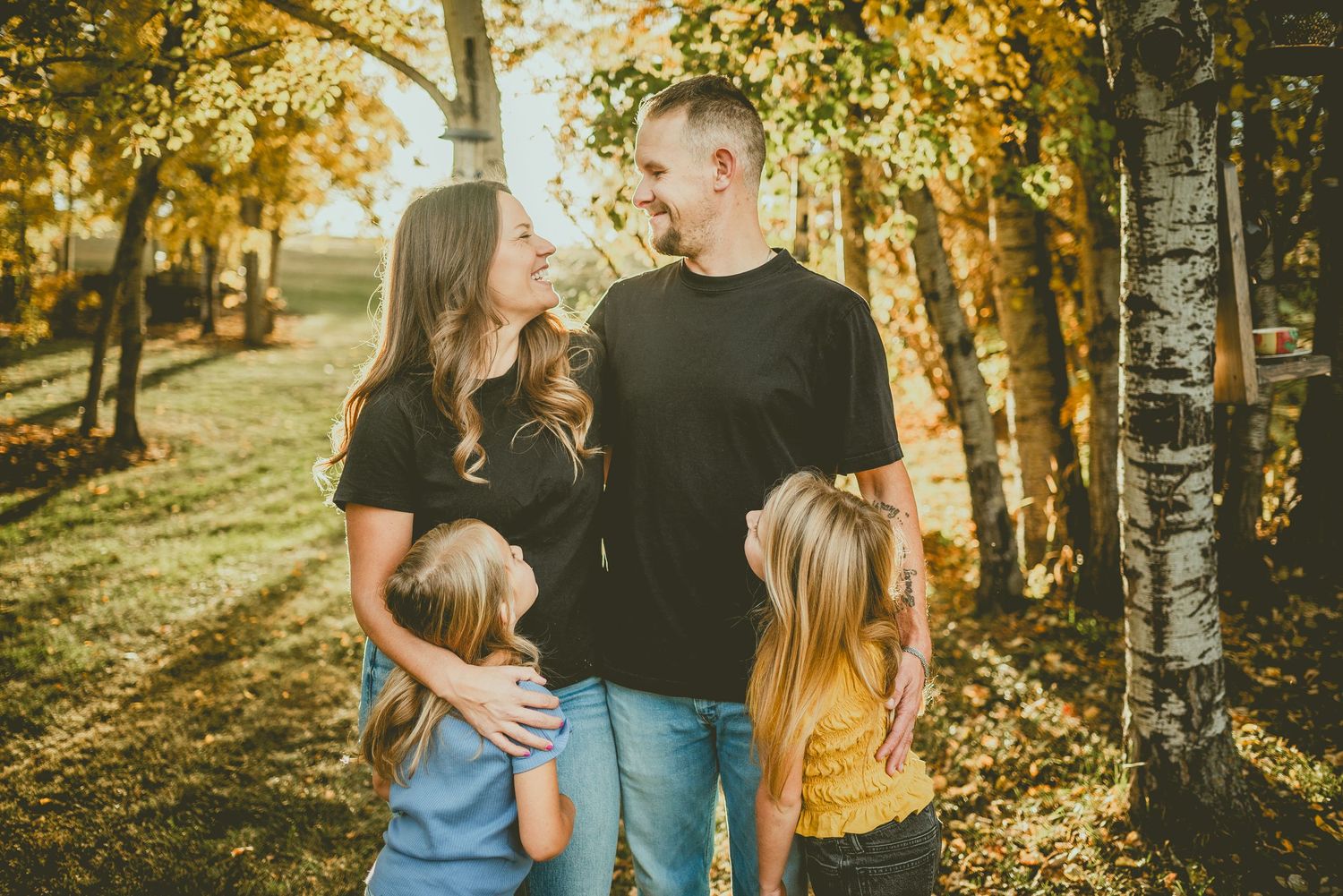 A family shares a tender moment together in a sunlit autumn forest with golden leaves and birch trees.