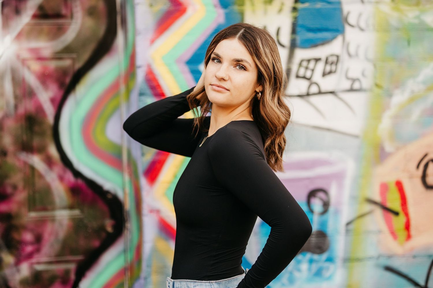 Young woman in black long-sleeve top poses confidently in front of colorful graffiti wall.